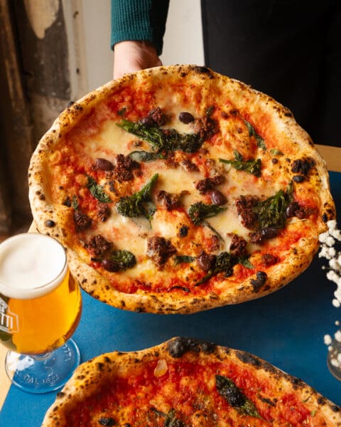 person holding neapolitan style pizza and a glass for beer on the side